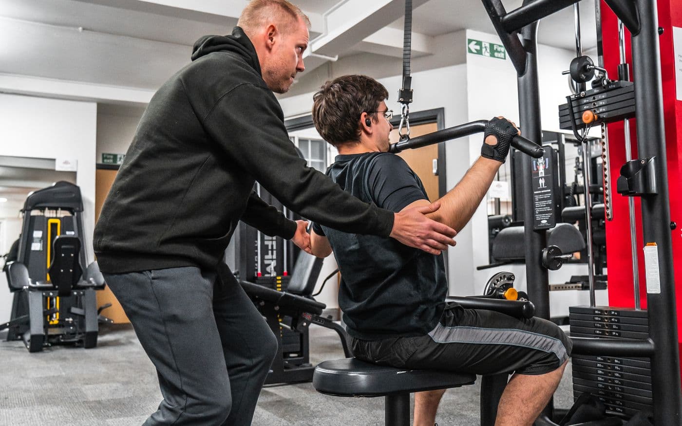 Trainer korrigiert einen Gast an einer Kraftmaschine im Fitnesscenter